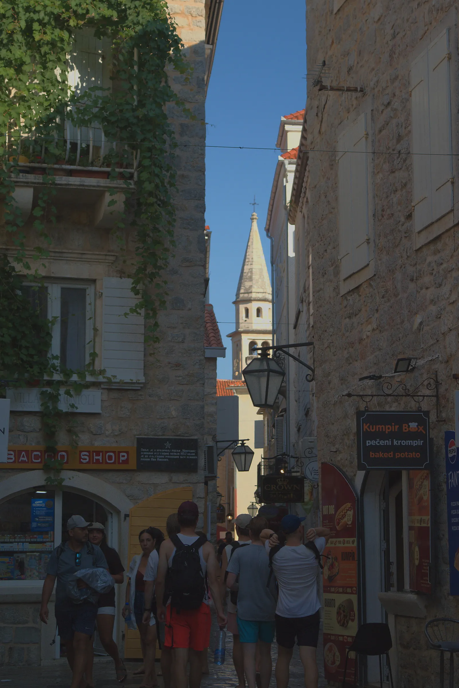 Un vicolo stretto della città vecchia di Budva, affollato di persone, con un campanile bianco sullo sfondo