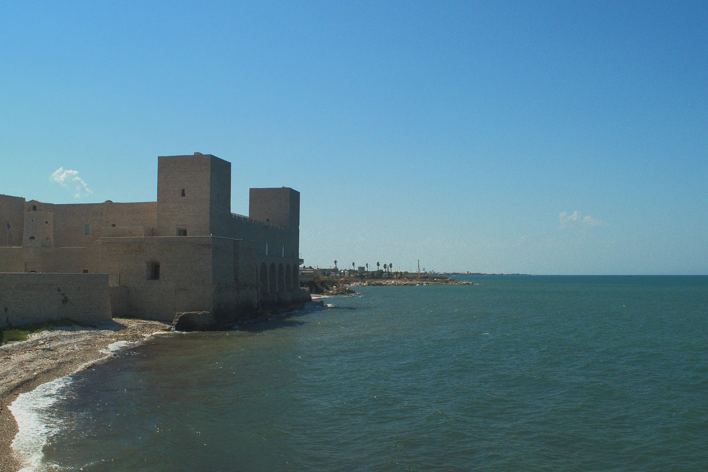 Il castello svevo di Trani affacciato sul mare, in controluce, con la spiaggia di ciottoli in primo piano